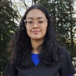 Lady with dark wavy hair. She is wearing a black scrub top with blue t-shirt.